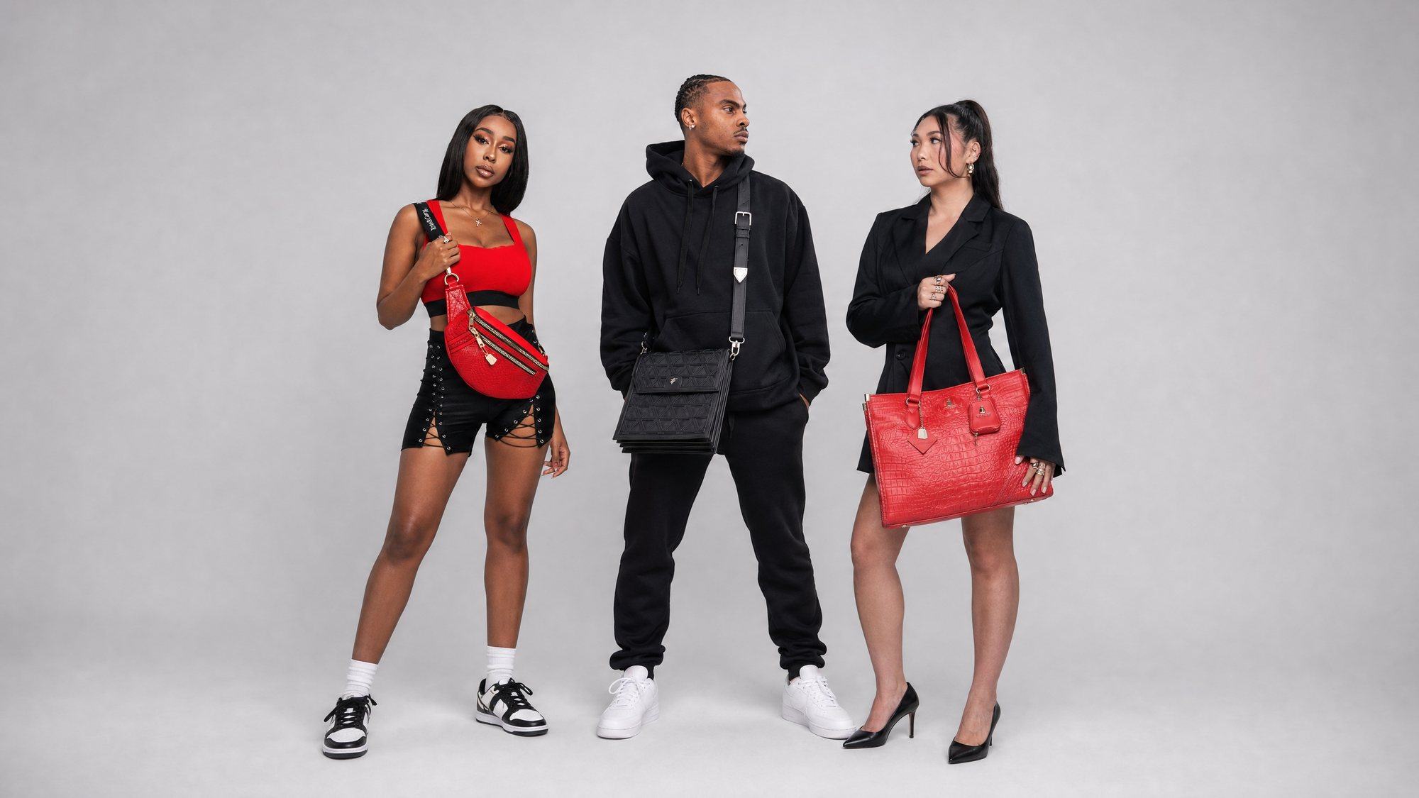 Three models in a grey studio carrying a red Apollo fanny pack, a black woven messenger bag and a red crocodile tote.